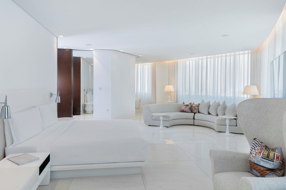 A modern hotel room with a large bed, a curved sofa, a small table, and a chair.