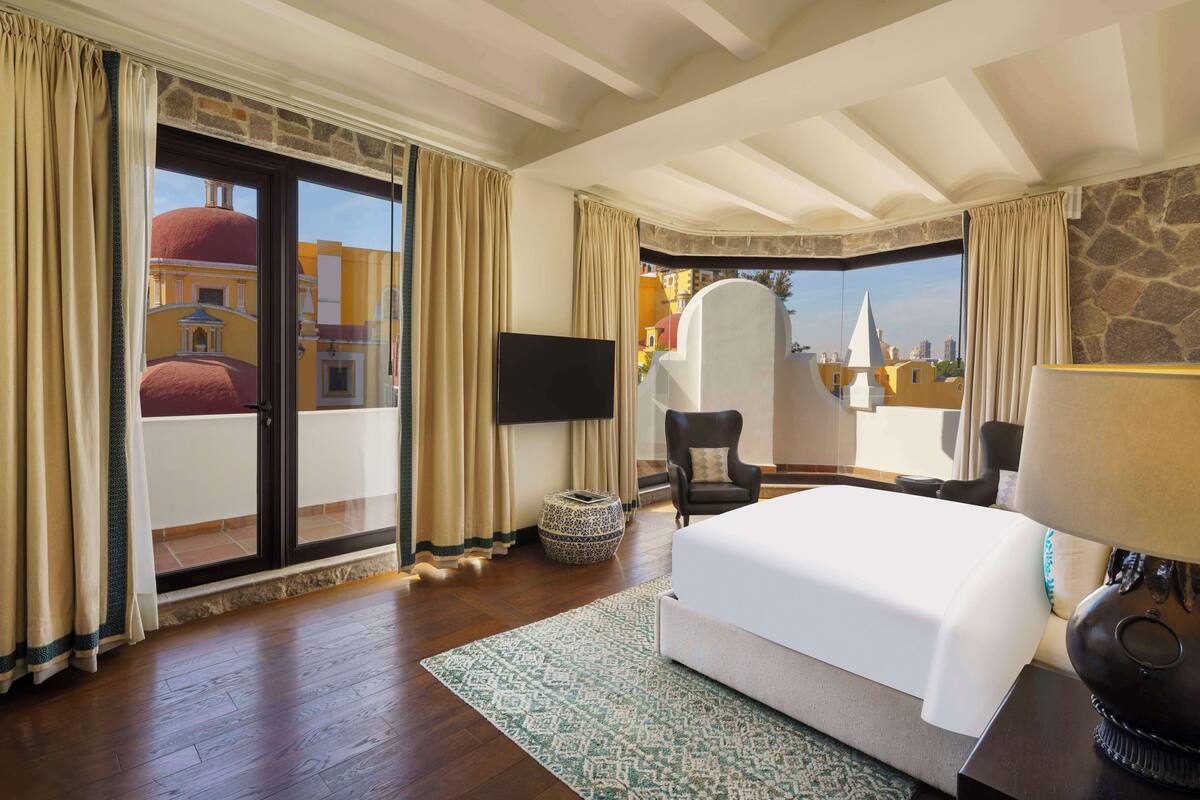 A hotel room with a large bed, a flat-screen TV, a chair, a lamp, and a view of buildings through a glass door.