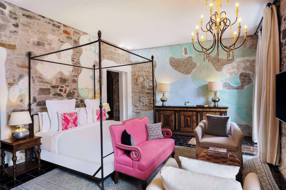 A four-poster bed with a pink upholstered headboard, a white sofa, a wooden cabinet, and a chandelier.