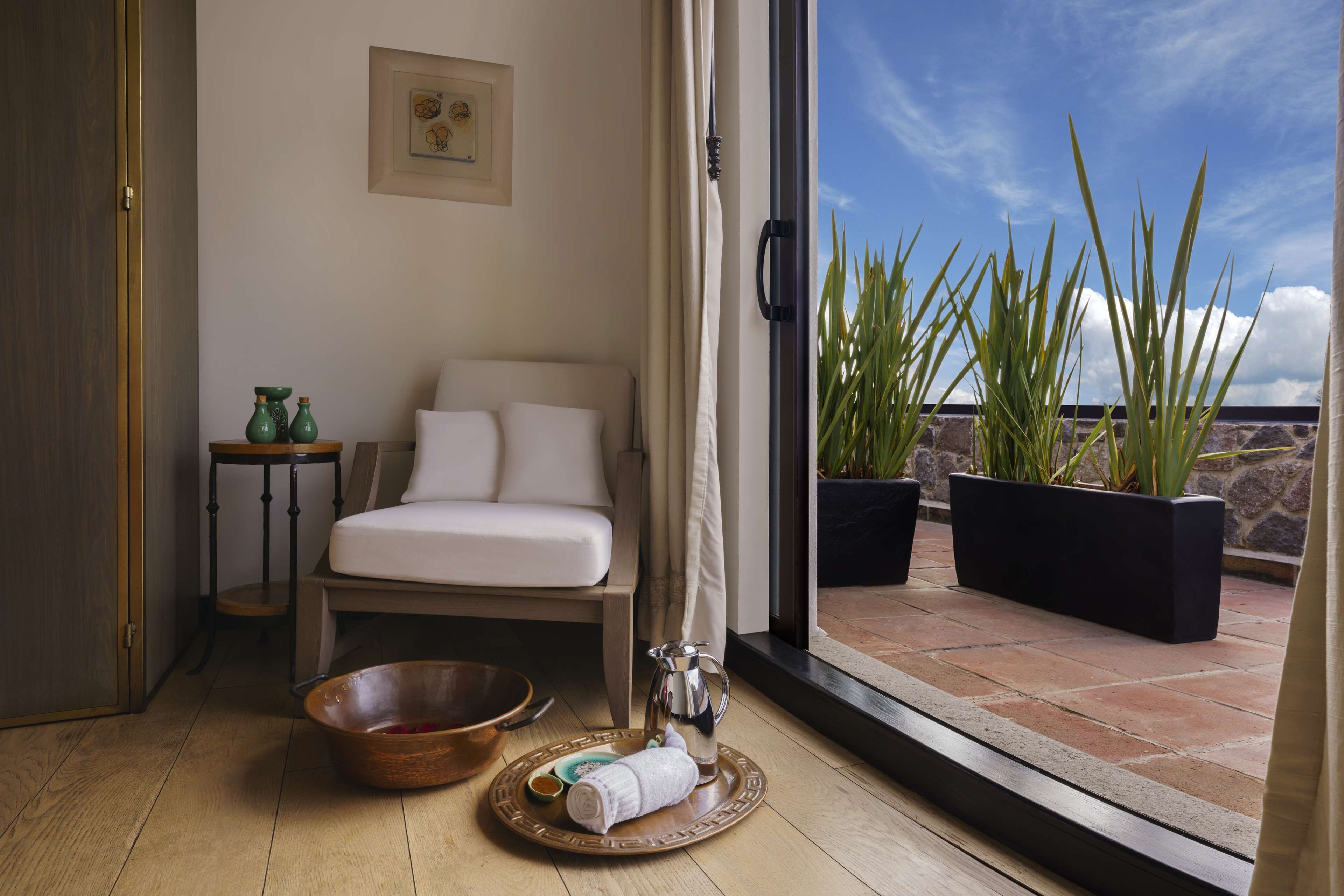A room with a bed, a small table with a bowl and a pitcher, a window with a view of plants and a clear sky.