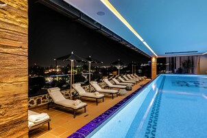 A poolside area with lounge chairs and umbrellas, a tiled pool, and a city view at night.