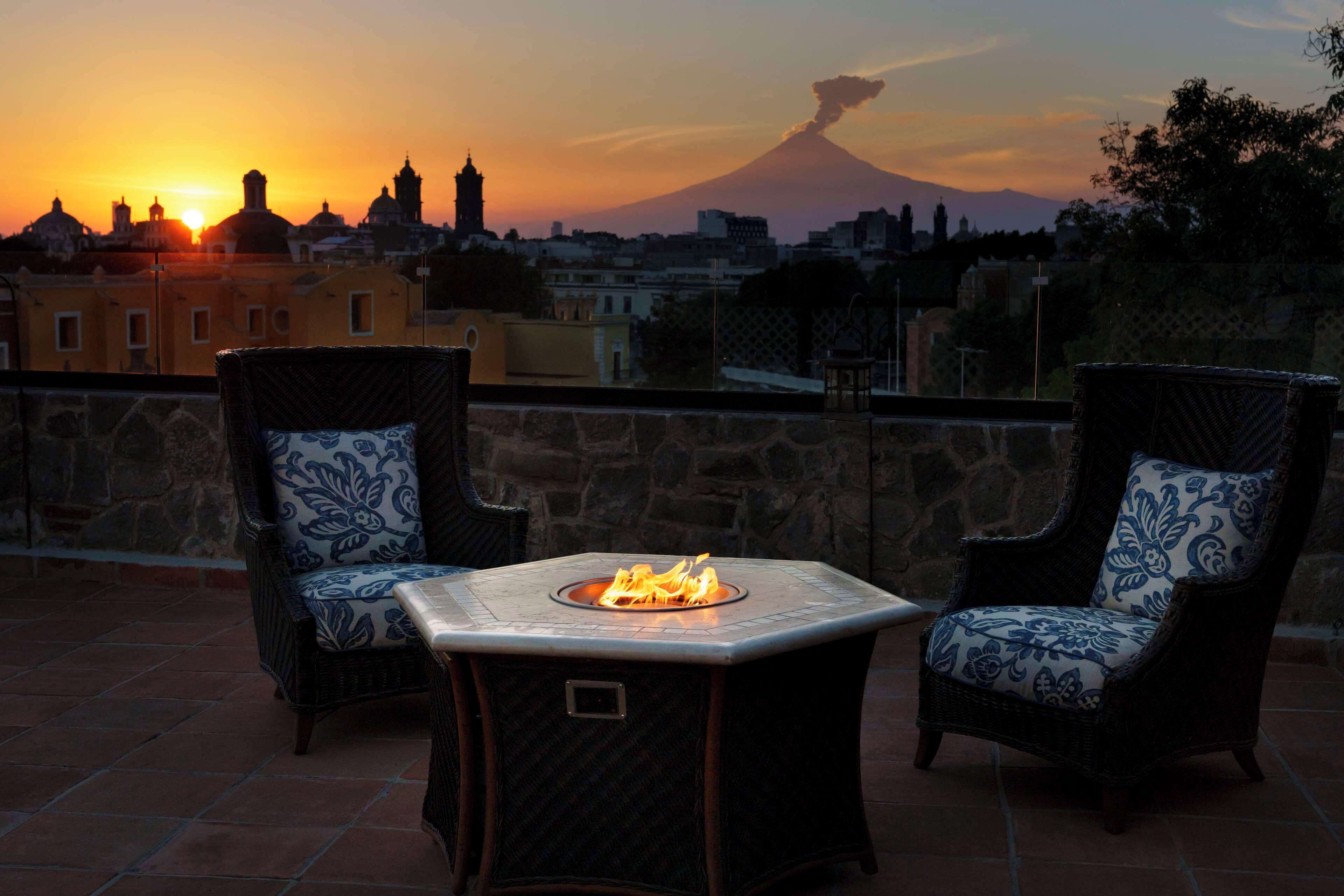 A terrace with two wicker chairs and a central fire feature, overlooking a cityscape and a volcano at sunset.