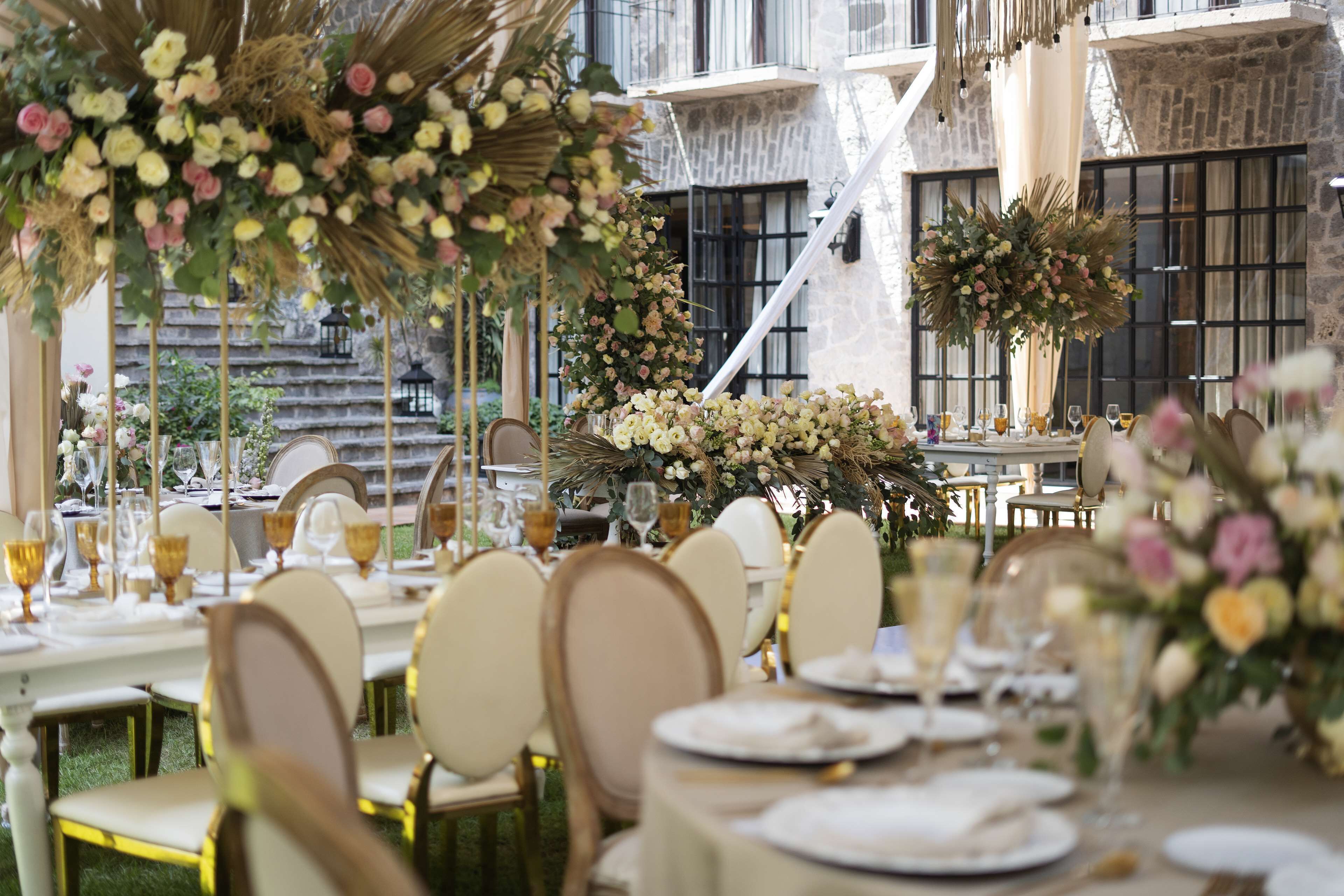 Un área de comedor al aire libre con mesas dispuestas para un evento formal, rodeada de abundantes arreglos florales y un edificio de piedra al fondo.