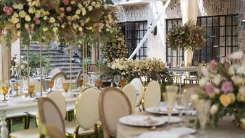 Un área de comedor al aire libre con mesas dispuestas para un evento formal, rodeada de abundantes arreglos florales y un edificio de piedra al fondo.