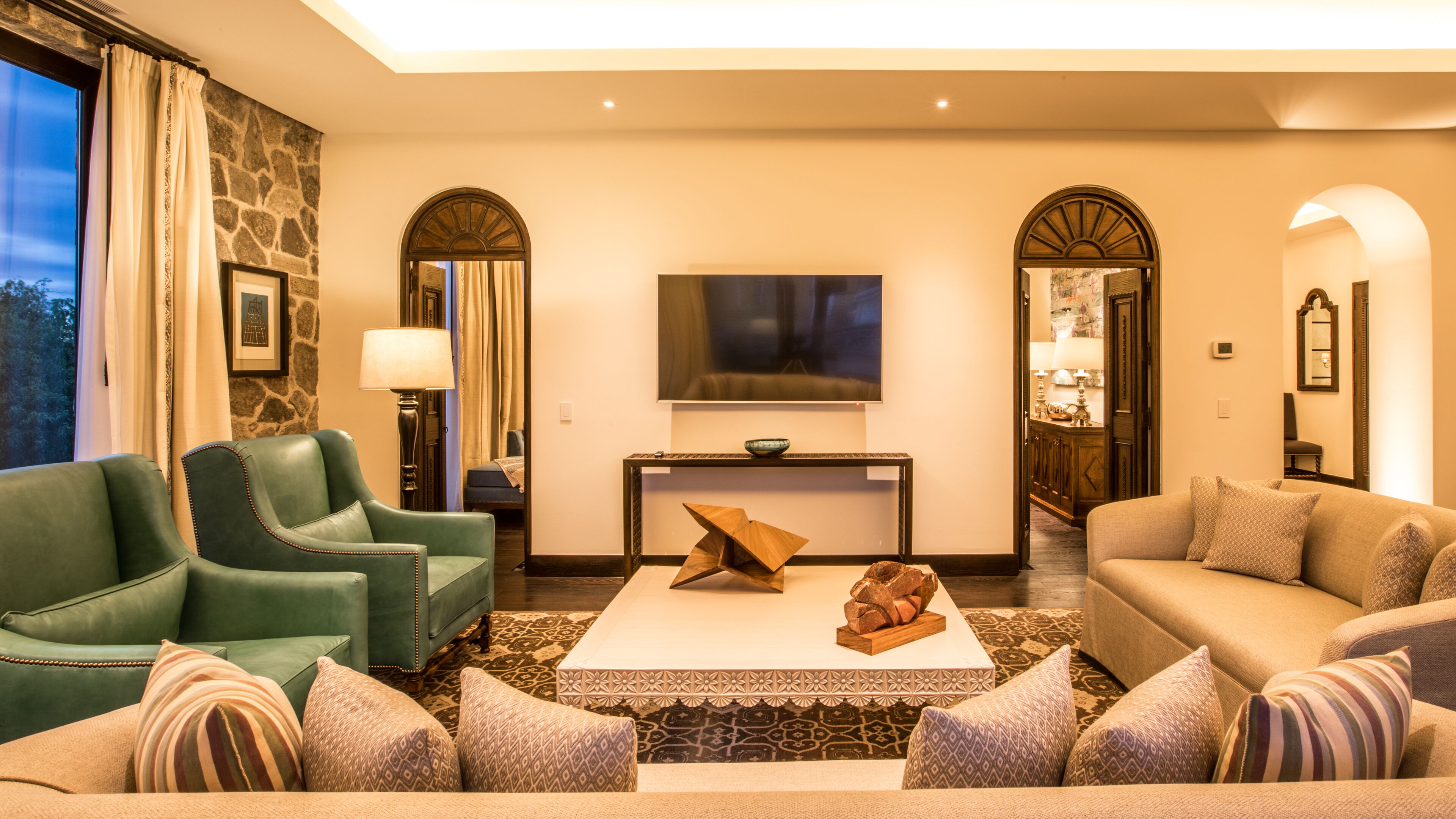 Una sala moderna con un sofá verde, un sofá beige y una mesa de centro con objetos decorativos. Hay un televisor montado en la pared, una lámpara y una pared de acento de piedra.