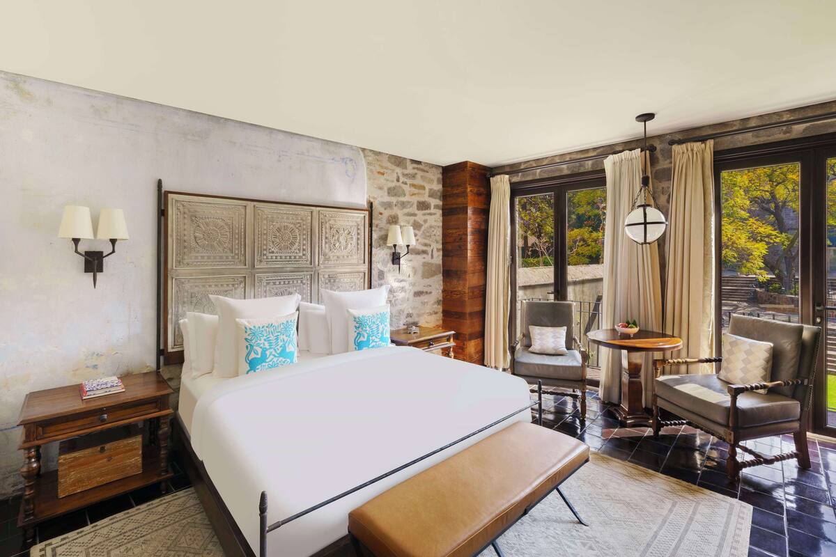 A hotel room with a large bed, a bench, a chair, a small table, and a view of the outdoors through large windows.