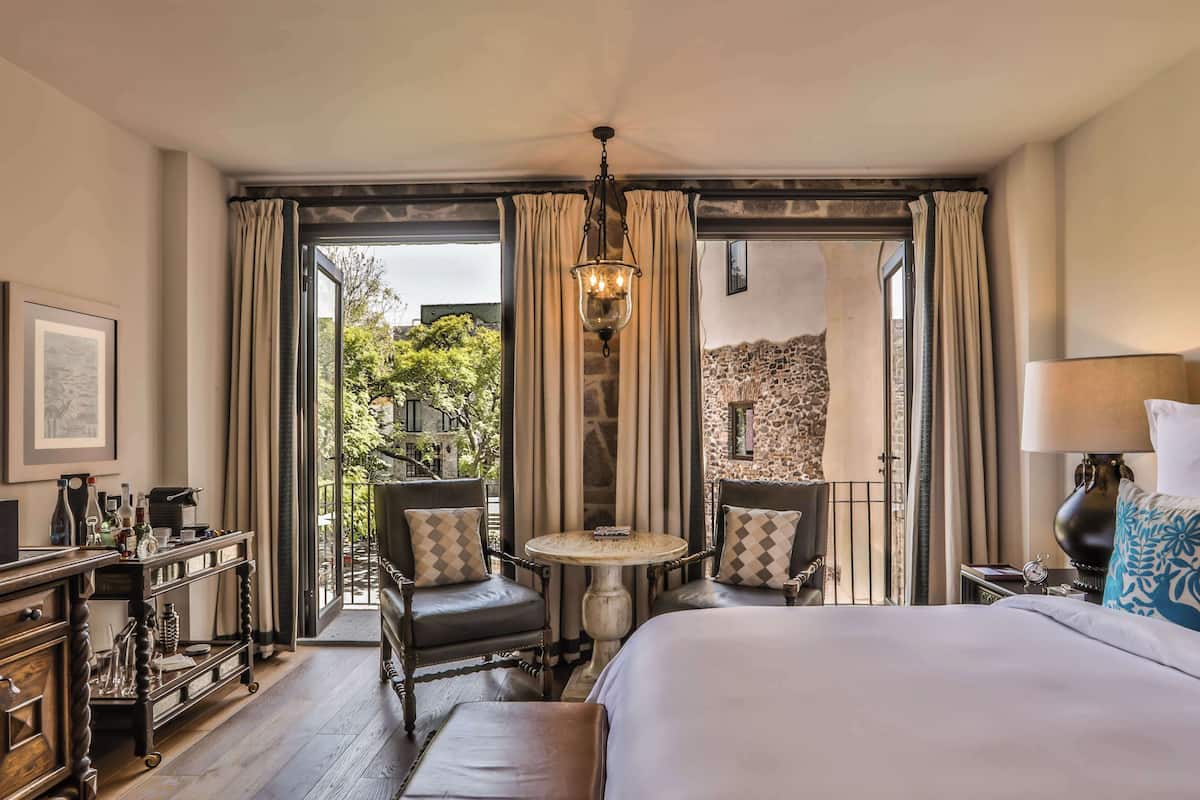 A hotel room with a bed, two armchairs, a small table, a chandelier, and a view of a stone wall and greenery outside.