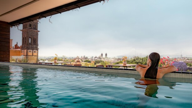 Una persona descansando en una piscina con vistas a una torre histórica y al paisaje urbano.
