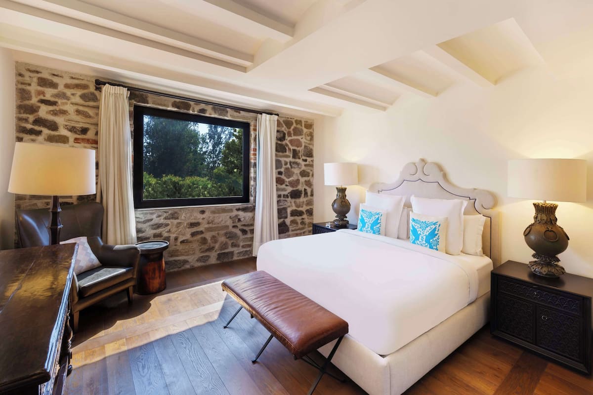 A bedroom with a large bed, a bench, a desk, and a stone wall.