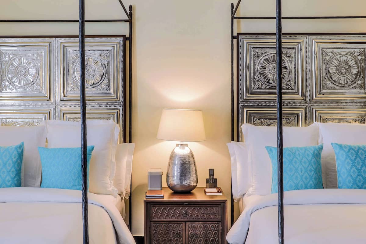 A hotel room with two beds, a nightstand with a lamp, and ornate headboard designs.