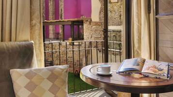 Una mesa redonda con un libro y una taza encima, una silla con un cojín de cuadros, y vista a un patio con un muro de piedra y un edificio morado.