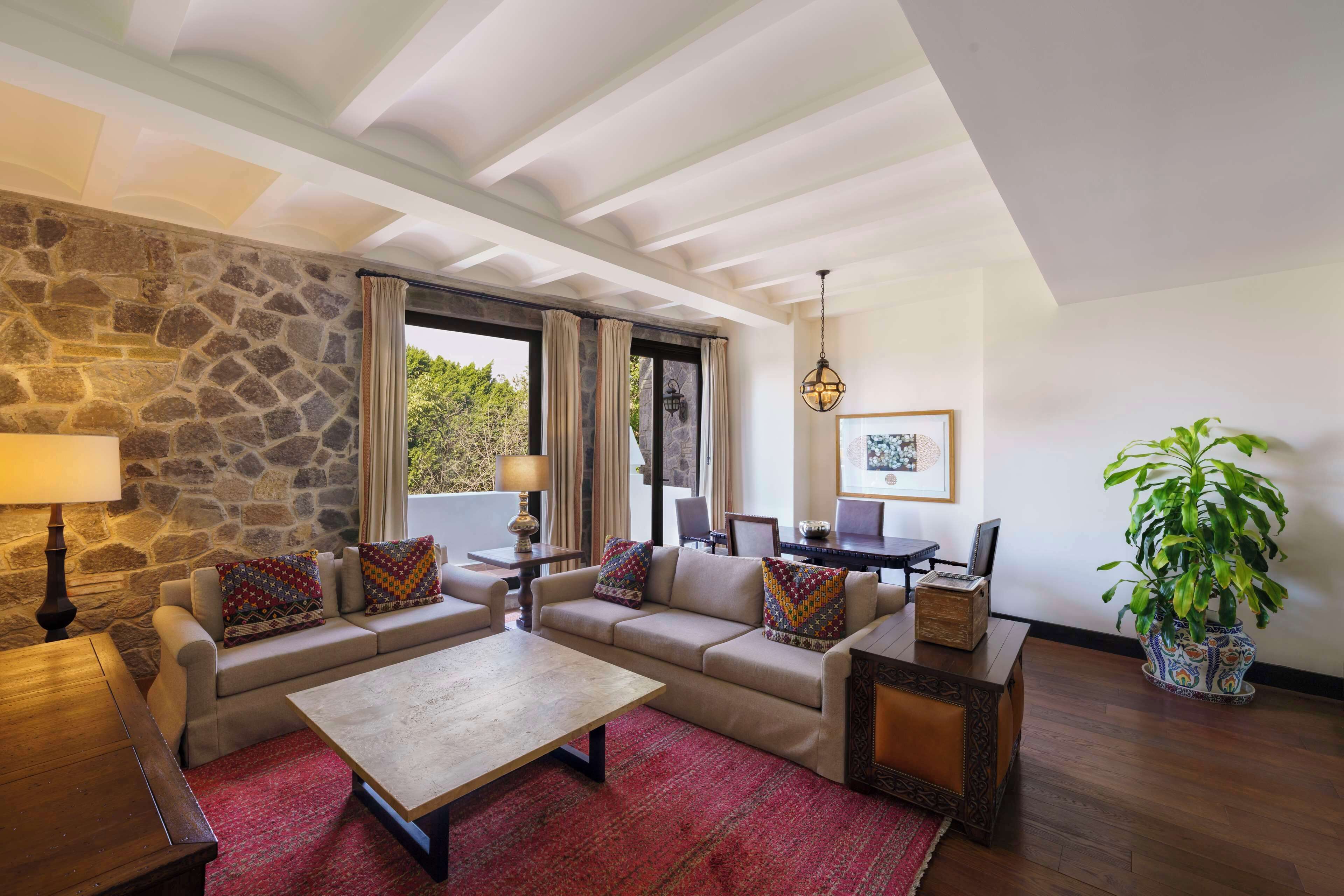Una sala de estar con pared de piedra, un conjunto de sofás, una mesa de centro, una mesa de madera, una lámpara, una planta en maceta y vistas a la vegetación exterior.