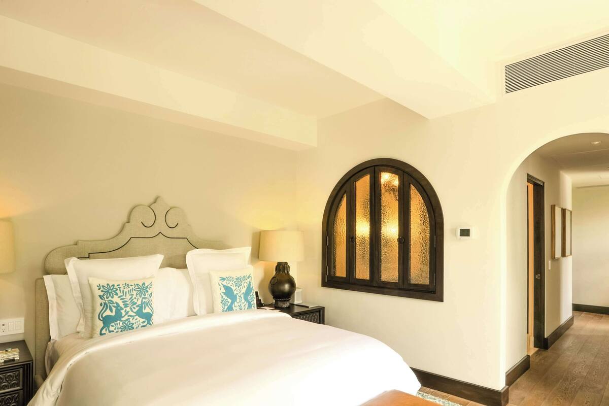 A bedroom with a large bed, a nightstand, a lamp, a decorative wall piece, and a hallway.