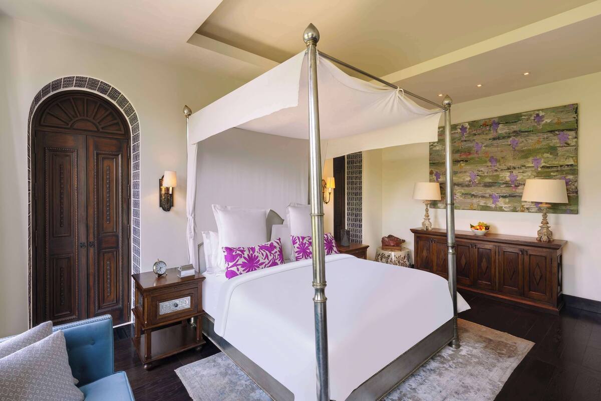A four-poster canopy bed with a nightstand and lamp, a wooden wardrobe, and a large floral painting on the wall.