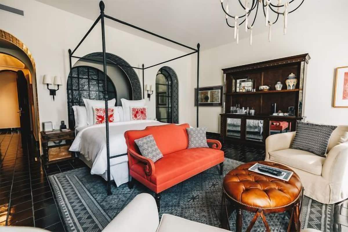 A four-poster bed, a red sofa, a white armchair, a wooden cabinet, and a patterned rug in a hotel room.