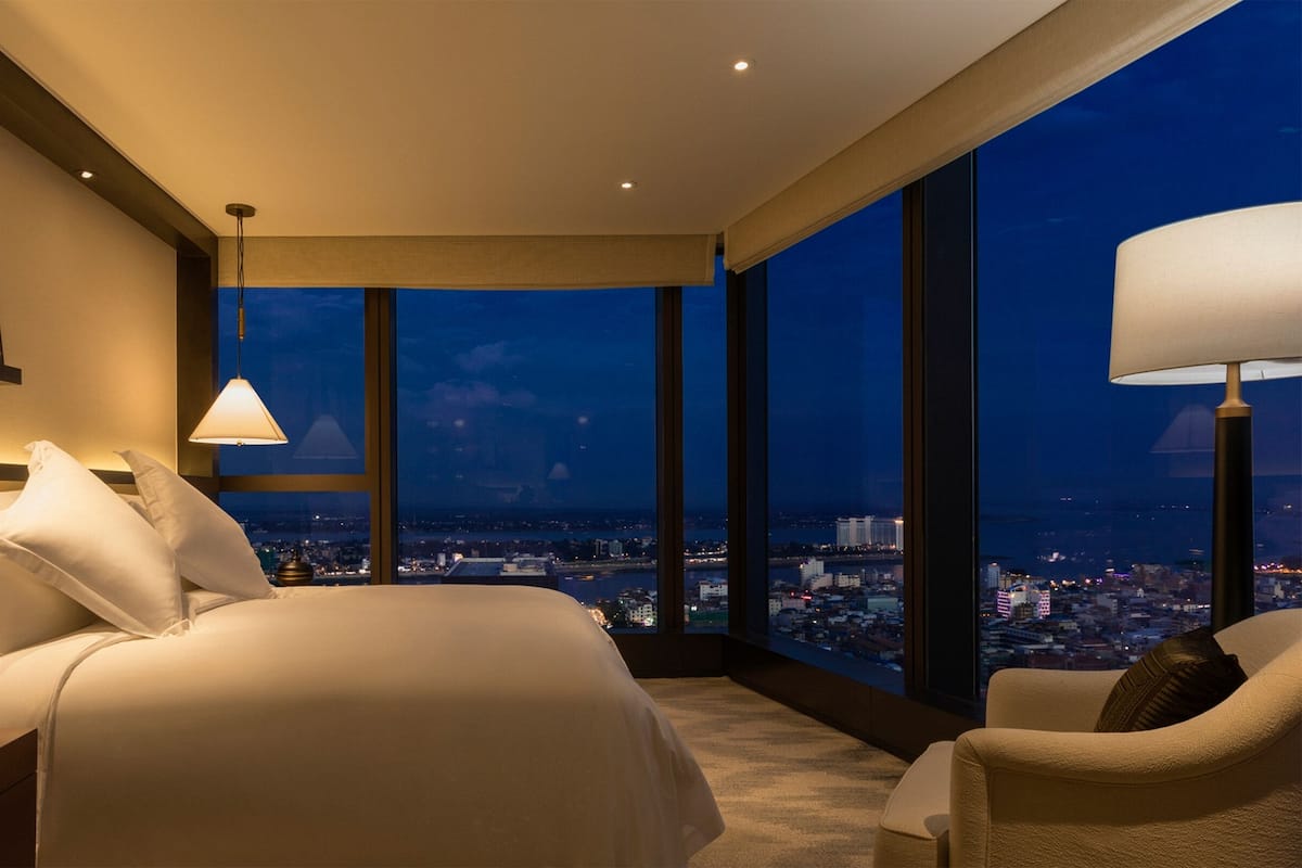 Une chambre d’hôtel avec un grand lit, une chaise, une table de chevet et une vue sur le paysage urbain grâce à de grandes fenêtres.