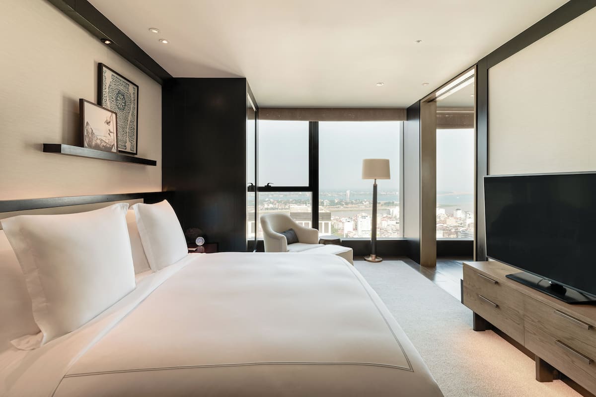 Une chambre d’hôtel moderne dotée d’un grand lit, d’un téléviseur à écran plat, d’un coin salon avec un fauteuil et une table, et d’une grande fenêtre donnant sur la ville.