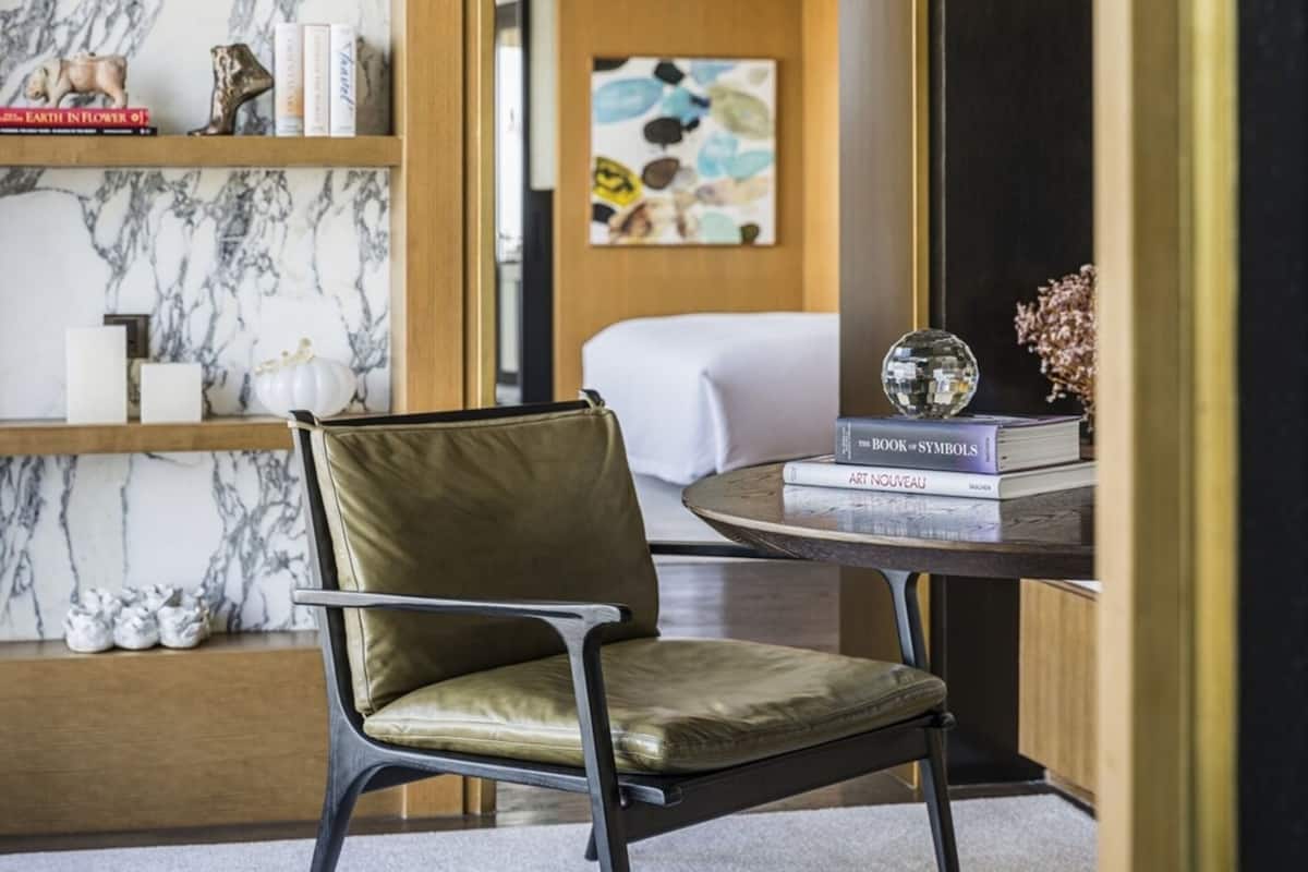 Une chambre moderne avec un fauteuil en cuir, une table ronde sur laquelle sont posés des livres, un mur en marbre et un lit à l’arrière-plan.