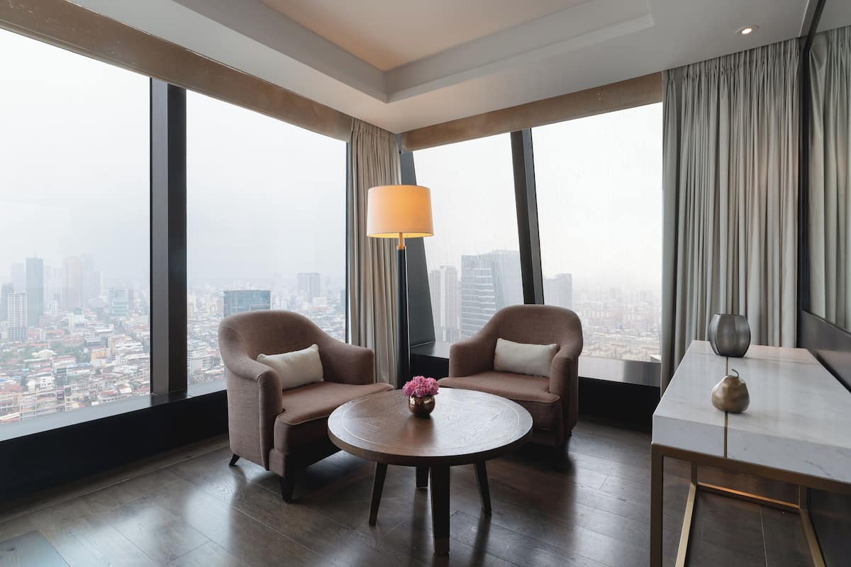 Une chambre d’hôtel moderne avec deux fauteuils, une table ronde, une lampe et une vue sur le paysage urbain.