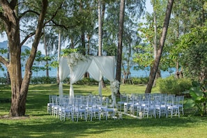An outdoor wedding setup with white chairs and draped fabric, surrounded by trees and a body of water.
