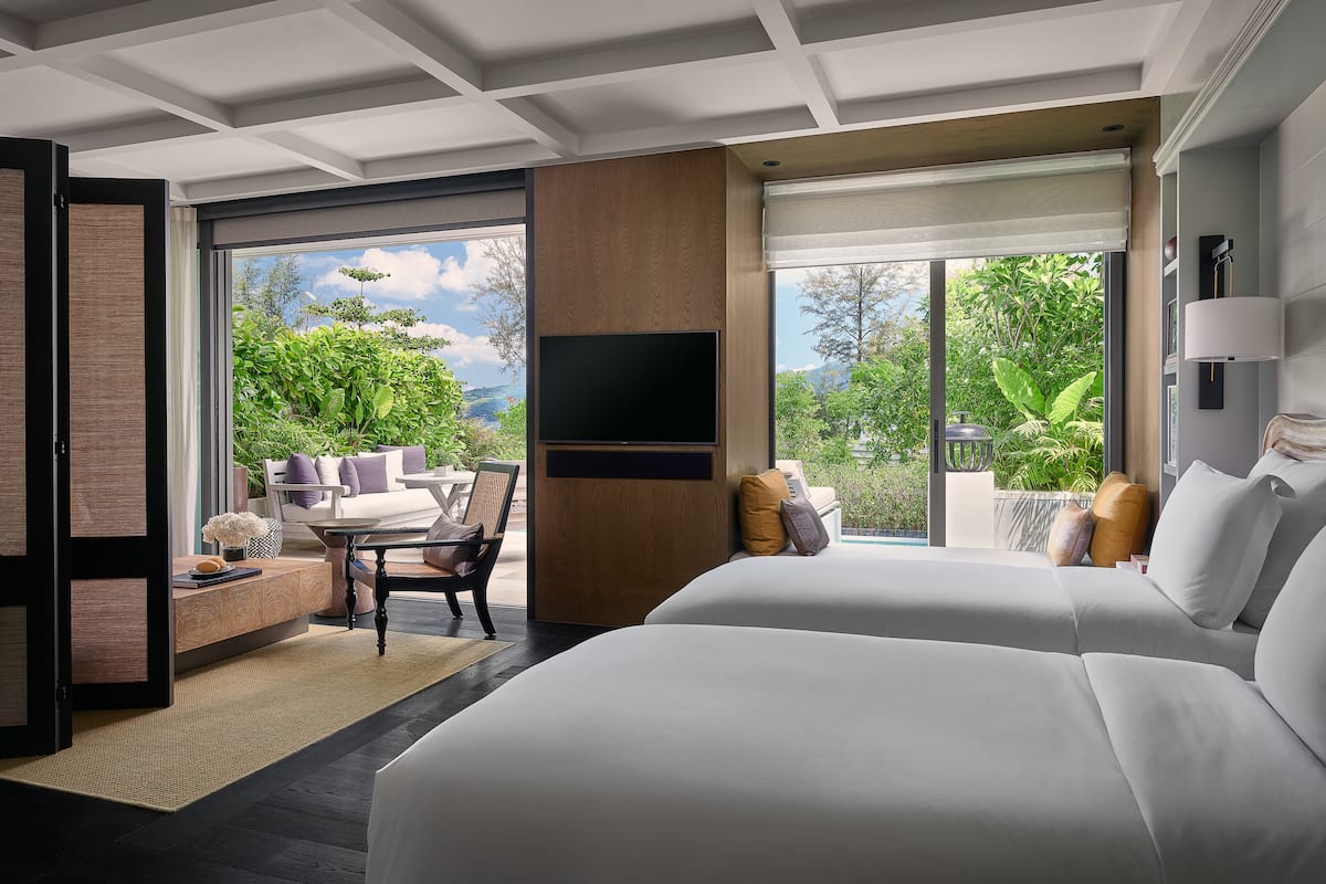 A modern hotel room with a large bed, a seating area with a table and chairs, a flat-screen TV mounted on the wall, and a balcony with a view of greenery and mountains.