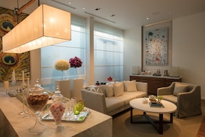 A modern living room with a sofa, armchairs, a coffee table, and a dining area with glassware and food.
