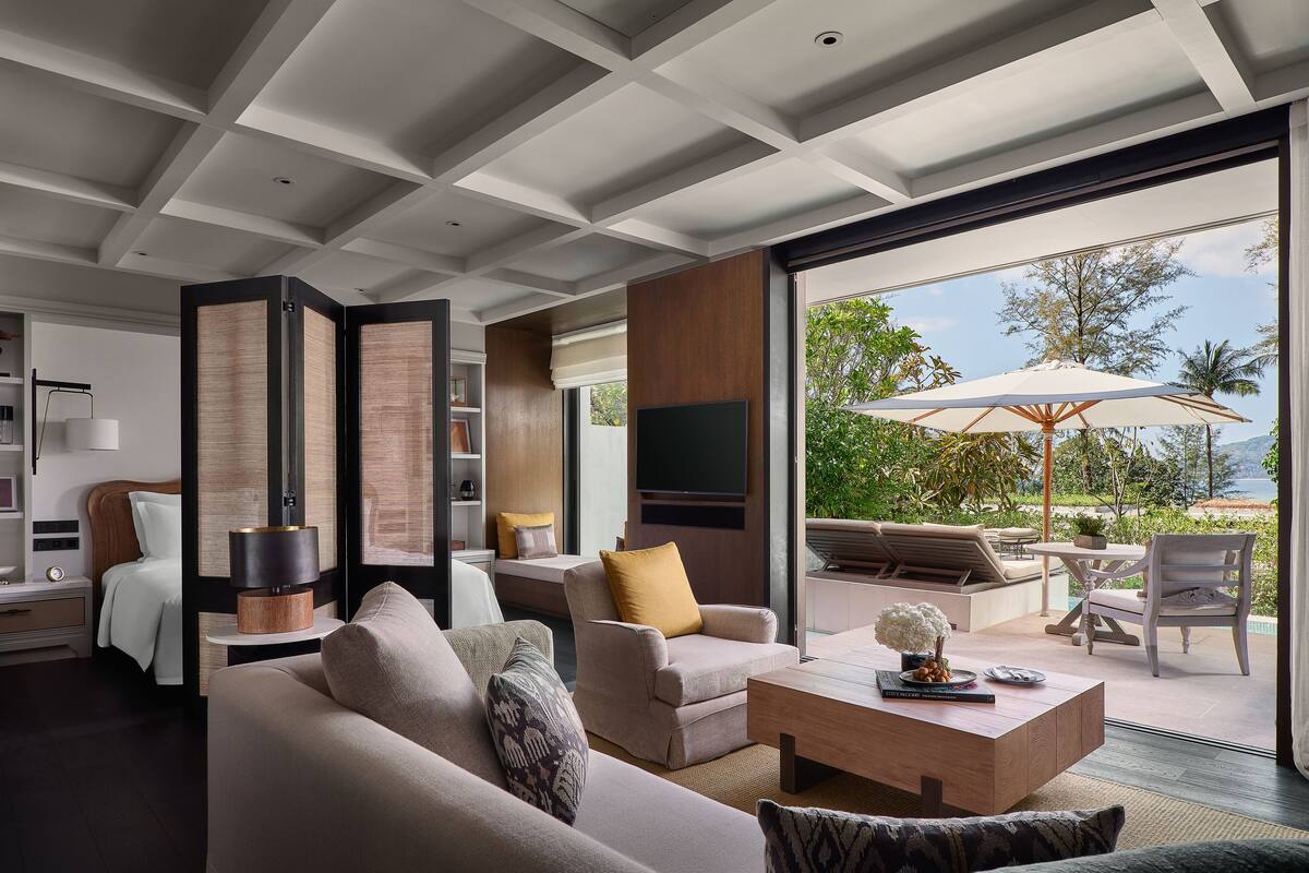A modern living room with a sofa, armchairs, a coffee table, and a TV. There is a large window with a view of the outdoors.