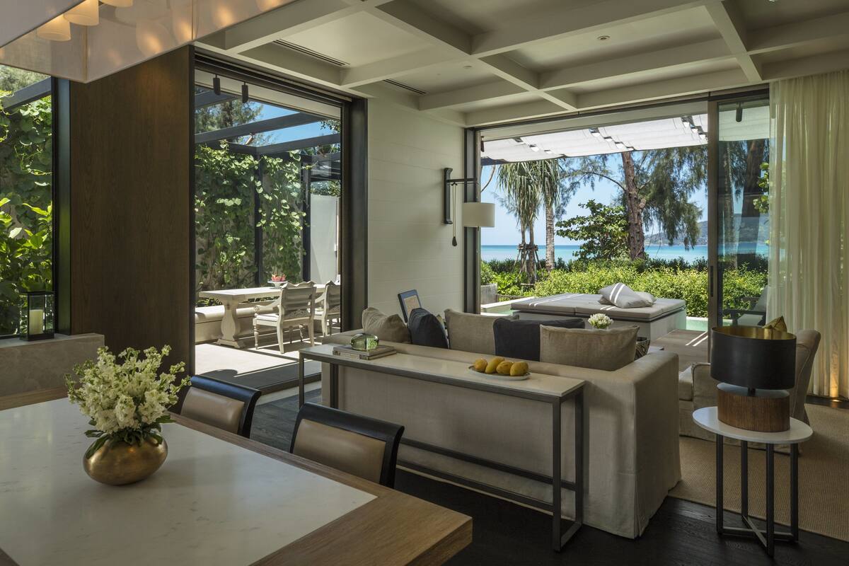 A modern living room with a sofa, coffee table, and dining area. Large windows offer a view of the outdoors.
