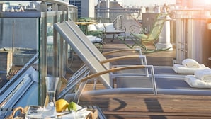 Um deck iluminado pelo sol, com cadeiras de lounge, uma mesa de vidro com bebidas e vista para o horizonte da cidade.