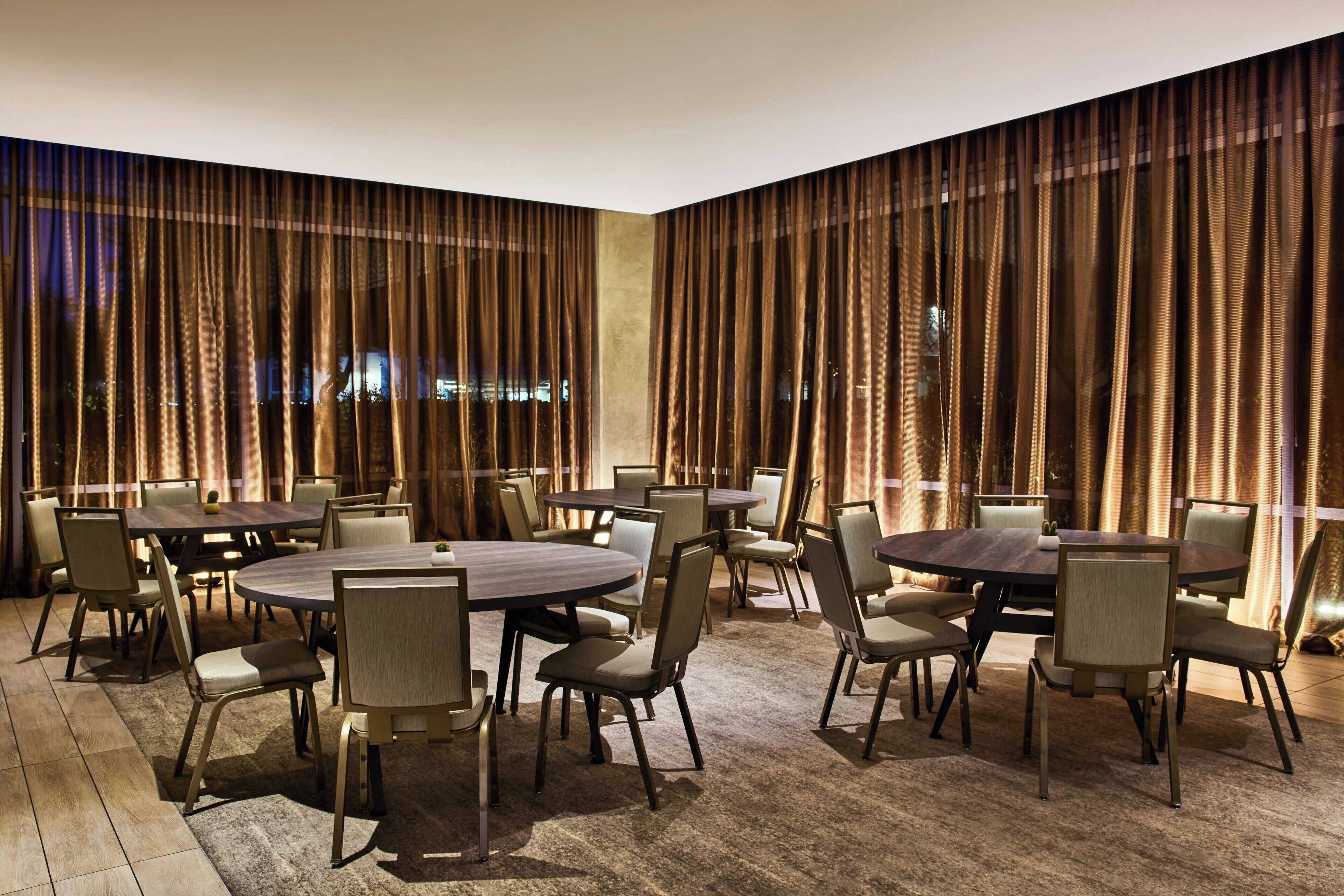 A conference room with multiple round tables and chairs, large windows with curtains, and a carpeted floor.