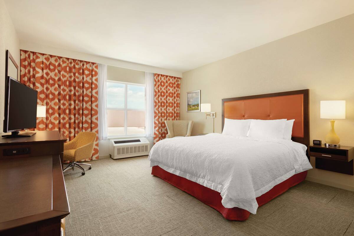A hotel room with a large bed, a desk with a television, a chair, and a window with curtains.