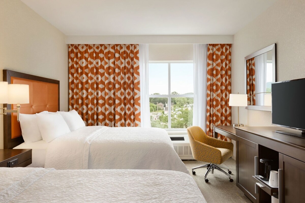 A hotel room with two beds, a desk with a TV, a chair, and a large window with curtains.