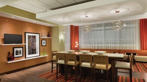 A hotel dining area with a long table, multiple chairs, and a large window with curtains. - Hampton Inn St. Albans (St. Albans)