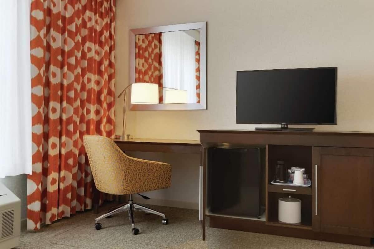 A hotel room with a desk, a television, a chair, a mirror, and a cabinet with a coffee maker.