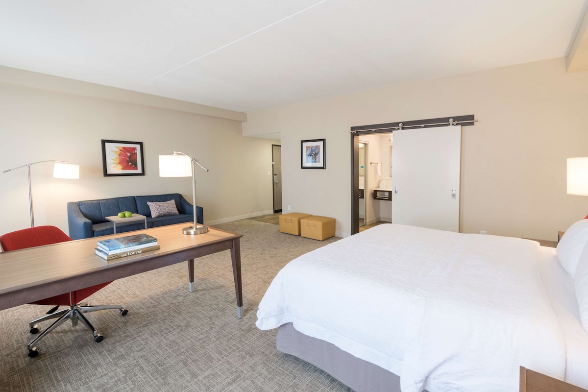 Ett hotellrum med en stor säng, ett skrivbord med en stol, en soffa, två inramade tavlor på väggen och en dörr som leder till ett badrum.