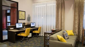 A hotel room with a desk, two computers, a yellow chair, a sofa with yellow cushions, and a decorative plant.