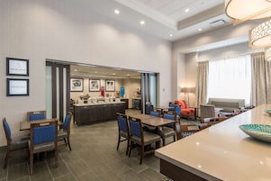 Un vestíbulo de hotel con mostrador de recepción, zona de comedor con mesas y sillas, y un área de descanso con sofá y sillones.