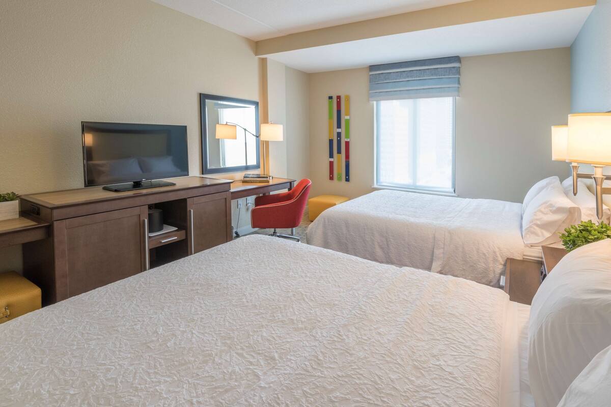 Ett hotellrum med två sängar, ett skrivbord med en tv, en röd stol och ett fönster med gardiner.