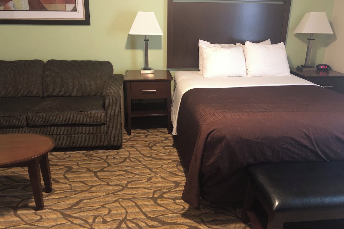 A hotel room with a bed, a sofa, a nightstand, a lamp, and a clock.