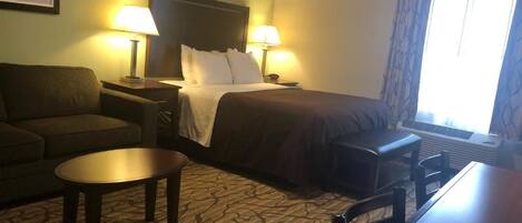 A hotel room with a bed, two bedside lamps, a sofa, a small table, and a window with curtains.