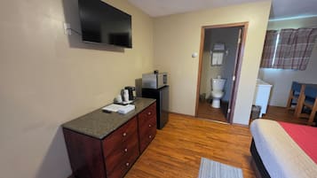A hotel room with a flat-screen TV mounted on the wall, a wooden dresser, a bed with a red blanket, a small kitchenette, and a bathroom visible through an open door.