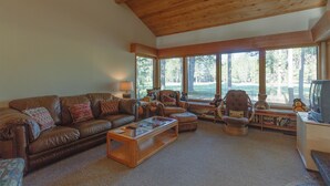 A spacious living room with a brown leather couch, a wooden coffee table, and a television on a stand. Large windows provide a view of the outdoors. - Augusta 4 (Bend)
