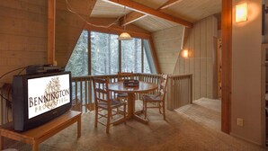 A cozy cabin interior with a wooden dining table and chairs, a television on a stand, and a large window offering a view of trees.