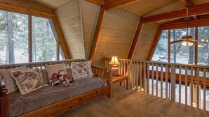 A cozy loft bedroom with a wooden ceiling, a bed with floral pillows, a nightstand, and a window overlooking a snowy landscape.
