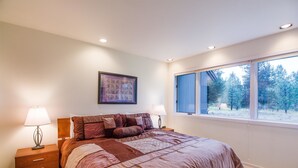 A bedroom with a large bed, two bedside tables with lamps, a window with a view of trees, and a framed picture on the wall. - Bachelor 5 (Bend)