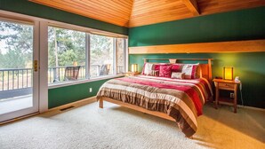 A bedroom with a large bed, a nightstand, a window with a view of trees, and a wooden ceiling. - Bachelor 5 (Bend)