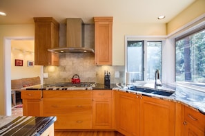 A kitchen with wooden cabinets, a stainless steel range hood, a granite countertop, a red kettle, and a window with a view of trees. - Bachelor 5 (Bend)