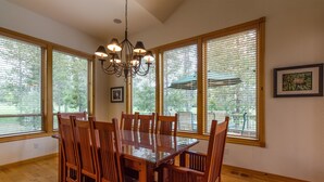 A dining area with a wooden table and chairs, large windows with blinds, a chandelier, and a framed picture on the wall.