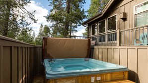 A hot tub in a wooden deck area with a wooden fence, wooden steps, and a view of trees and a building in the background.