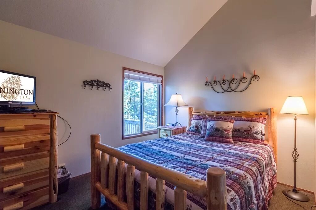 A bedroom with a wooden bed, a desk with a computer, a window with a view of trees, and a wall-mounted lamp.